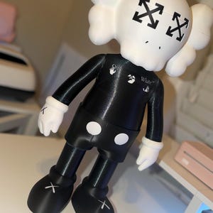 May include: A black and white figurine with a skull-shaped head and crossed-out eyes. The figurine has a black body, shorts with white circles, and white gloves. The feet have white crosses. The word "White" is printed on the chest.