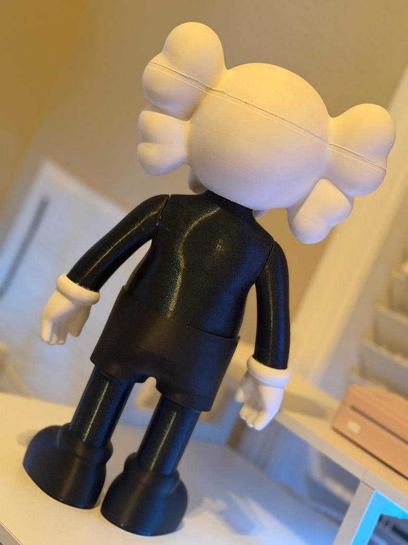 May include: A black and white figurine of a cartoon character with a rounded head and crossbones on the back. The figure is wearing a black outfit with white gloves and shoes. The figurine is standing on a white surface.