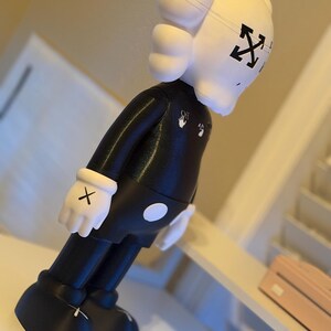 May include: A black and white figurine with a unique design. The figure has a white head with black cross-shaped eyes and a black body. The figure is wearing white gloves with black X's on the palms. The figure has the Off-White logo on the chest.