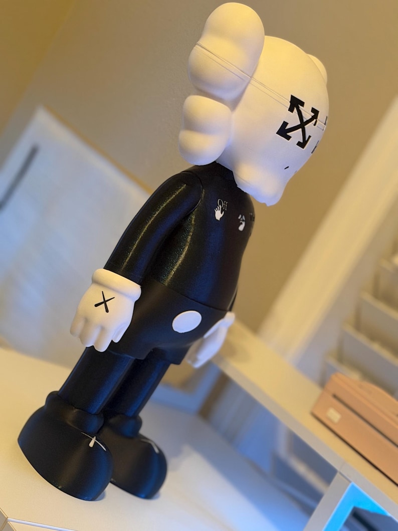 May include: A black and white figurine with a unique design. The figure has a white head with black cross-shaped eyes and a black body. The figure is wearing white gloves with black X's on the palms. The figure has the Off-White logo on the chest.