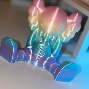 May include: A colorful figurine in a seated position, featuring a gradient of pink, yellow, blue, and purple hues. The figure has crossed-out eyes and is holding a money bag with a dollar sign. The figurine is a unique decorative item.