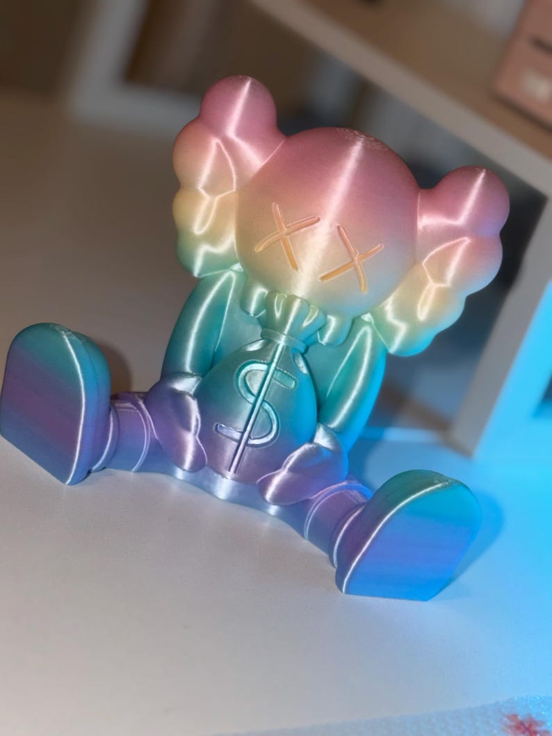 May include: A colorful figurine in a seated position, featuring a gradient of pink, yellow, blue, and purple hues. The figure has crossed-out eyes and is holding a money bag with a dollar sign. The figurine is a unique decorative item.