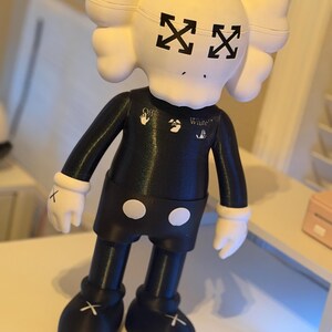 May include: A black and white figurine with a unique design. The figurine has a white head with black arrow-shaped eyes and a black body. The arms and legs are black, with white gloves and shoes. The figurine has the word "White" printed on the chest.