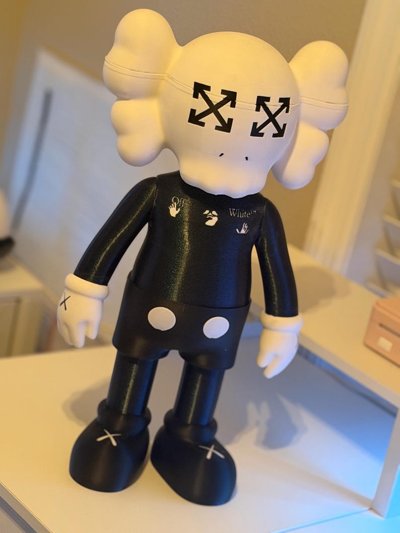 May include: A black and white figurine with a unique design. The figurine has a white head with black arrow-shaped eyes and a black body. The arms and legs are black, with white gloves and shoes. The figurine has the word "White" printed on the chest.