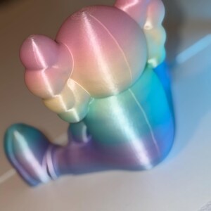 May include: A colorful, gradient-colored figurine in shades of pink, yellow, blue, and purple. The figurine has a rounded head with large ears and a seated pose. The surface has visible layer lines from the 3D printing process.