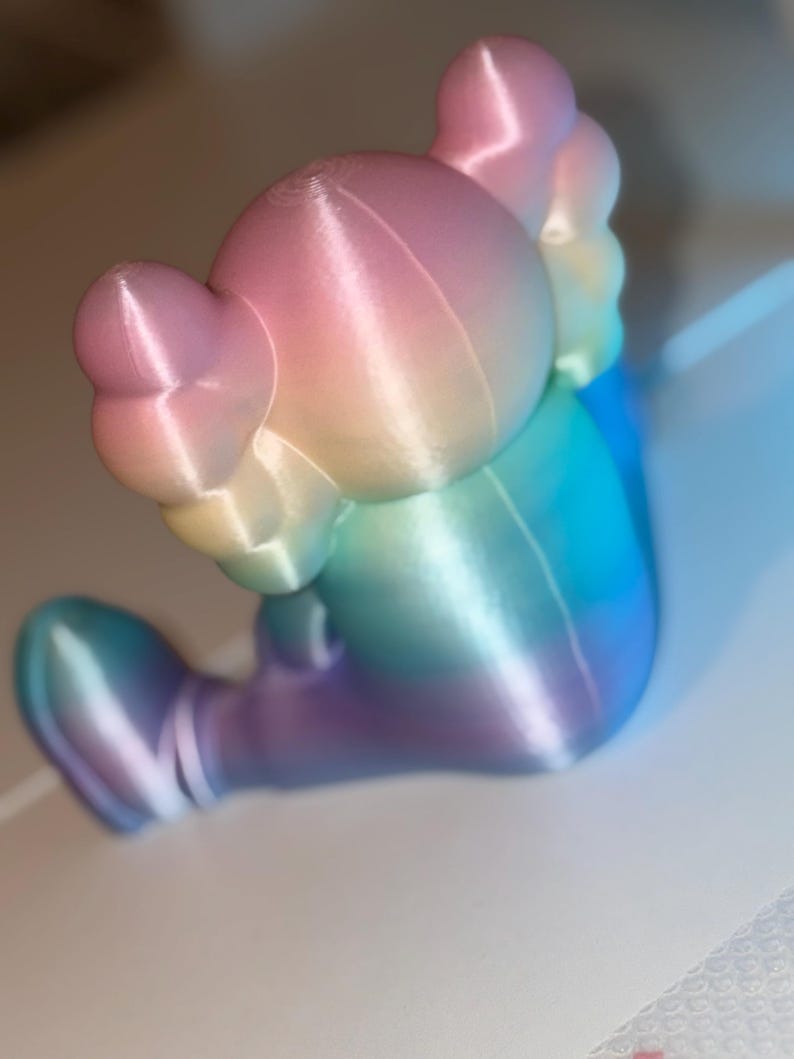 May include: A colorful, gradient-colored figurine in shades of pink, yellow, blue, and purple. The figurine has a rounded head with large ears and a seated pose. The surface has visible layer lines from the 3D printing process.