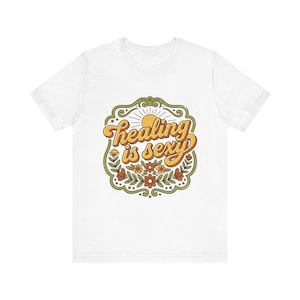 May include: White t-shirt with the text "Healing is Sexy" in a retro font. The design features a sun, flowers, and a decorative border in shades of yellow, orange, green, and brown.