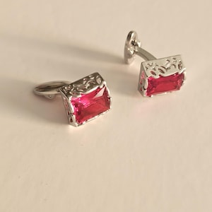 May include: A pair of silver-toned cufflinks featuring rectangular, faceted, vibrant red stones set within intricate, openwork metal frames. Each cufflink has a polished silver finish and a hinged toggle mechanism on the back for secure fastening.