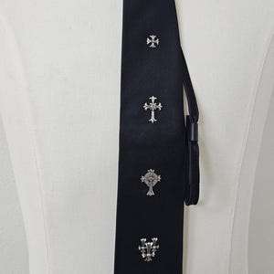 Può includere: Una cravatta nera con una trama a coste diagonali e cinque emblemi a croce in metallo color argento disposti verticalmente lungo il centro. La cravatta è annodata nella parte superiore e presenta un bordo inferiore a punta con finitura scura.
