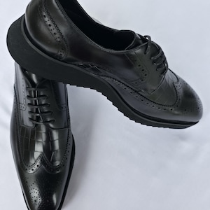 May include: A pair of black leather oxford shoes featuring a wingtip design with brogue detailing and a crocodile-embossed texture on the toe and side panels. The shoes have black laces and a thick, solid black platform sole with a ridged tread.