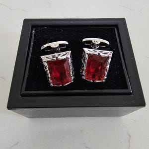 Puede incluir: Un par de gemelos plateados con piedras rectangulares facetadas de color rojo intenso engastadas en marcos de metal ornamentados. Los gemelos se presentan dentro de una caja negra cuadrada con un interior forrado de terciopelo.