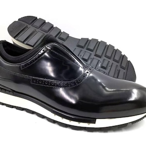 May include: Black slip-on shoe with a glossy finish, white sole, and a textured black rubber outsole. The shoe features decorative perforations and a black elastic collar. The second shoe is visible in the background.