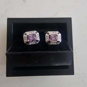 Puede incluir: Un par de gemelos plateados con una piedra preciosa rectangular morada en el centro, rodeada de piedras transparentes más pequeñas y detalles en esmalte morado. Los gemelos se presentan en una caja negra con interior de terciopelo.