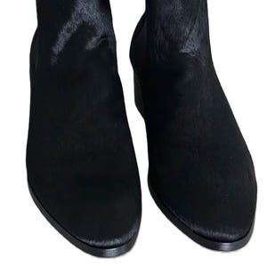 May include: A pair of black ankle boots with a textured, fur-like appearance. The boots have a low heel and a pull tab at the top. The footwear is set against a white background.