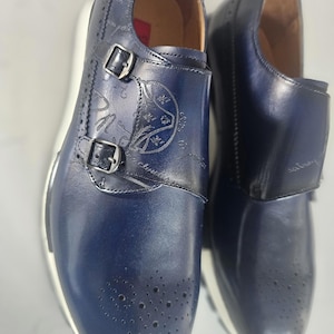 May include: A pair of navy blue leather double monk strap shoes featuring a decorative brogue toe cap, silver-toned metal buckles, and a subtle script pattern printed across the upper surface, set upon a thick white rubber sole.
