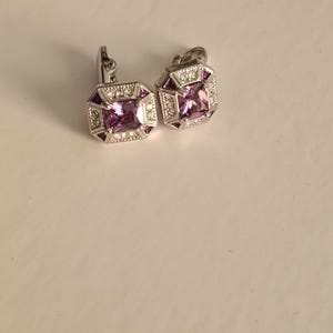 May include: A pair of silver-toned earrings featuring a central square-cut purple gemstone surrounded by a border of small clear stones and four triangular purple accents set within an octagonal frame with a hinged post backing mechanism.