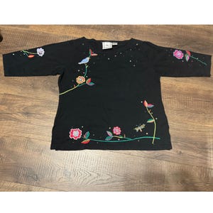 May include: Black three-quarter sleeve shirt with embroidered floral and bird design. The embroidery features pink, red, green, and yellow accents. A blue bird is perched on a branch.