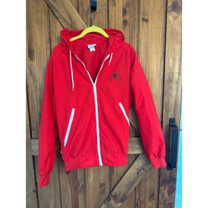 May include: A bright red hooded jacket with a white zipper and drawstrings. The jacket has two front pockets with white accents and a small black logo on the chest. The jacket is hanging on a yellow hanger.