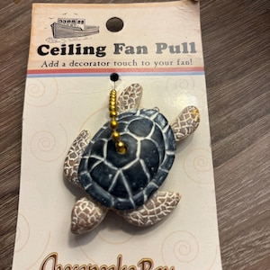 May include: A turtle-shaped ceiling fan pull with a dark grey shell and light brown textured limbs, attached to a gold-coloured ball chain. The item is mounted on a branded display card featuring a boat illustration and the text Chesapeake Bay LTD.