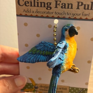 May include: A ceiling fan pull featuring a blue and yellow parrot figurine with green wing accents, attached to a gold-toned metal ball chain. The parrot is mounted on a white display card with gold polka dots and the text Ceiling Fan Pull.