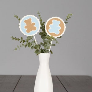 May include: Two brown and blue paper bear cutouts with scalloped edges are attached to sticks and placed in a white vase with green eucalyptus leaves.