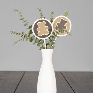 May include: A white vase with green eucalyptus leaves and two brown paper circles with brown bear silhouettes. The circles are attached to sticks and are arranged in the vase.