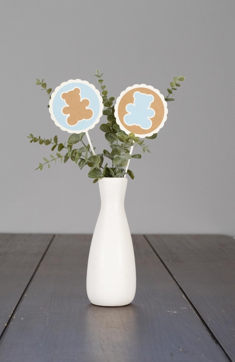 May include: Two brown and blue paper bear cutouts with scalloped edges are attached to sticks and placed in a white vase with green eucalyptus leaves.