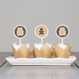 May include: Three brown cupcake liners with white cupcake wrappers, each topped with a brown teddy bear cupcake topper on a white rectangular plate.
