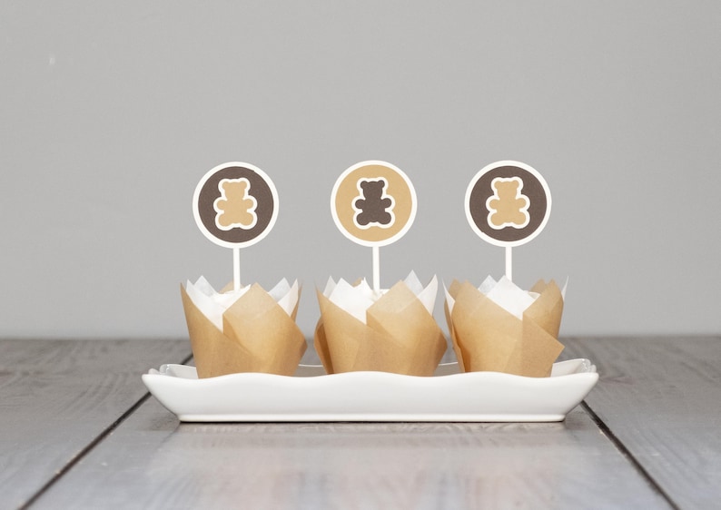 May include: Three brown cupcake liners with white cupcake wrappers, each topped with a brown teddy bear cupcake topper on a white rectangular plate.