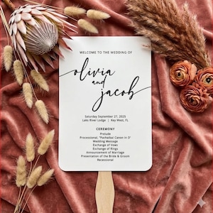 May include: A white wedding fan with the names "Olivia and Jacob" in elegant script. The fan includes the wedding date, location, and ceremony details. The background features dried flowers and a velvet fabric.