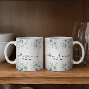 Pode incluir: Duas canecas de cerâmica branca com um padrão floral em aguarela em tons de azul e verde. Uma caneca apresenta o texto "Mr. James" e "05.11.2025" em letra preta, enquanto a outra apresenta "Mrs. James" e "05.11.2025".