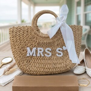 May include: A woven straw tote bag with the letters "MRS S" embellished with pearls. A white satin ribbon is tied to the handle. The bag is next to a pair of white shoes with pearl accents, a small mirror, and a gold bracelet.