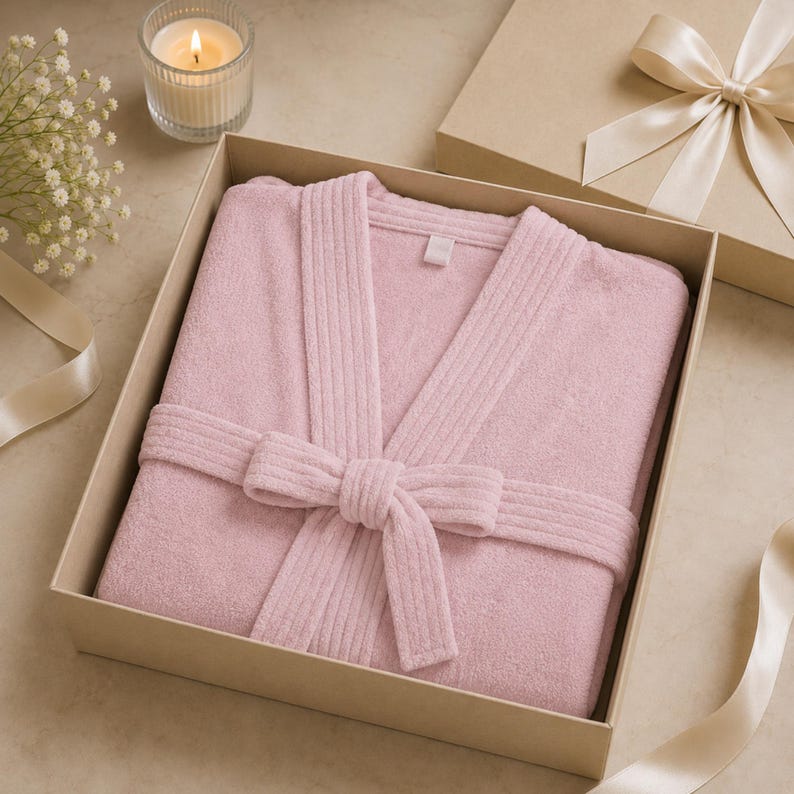 Pode incluir: Um roup&atilde;o de banho macio em rosa claro com gola canelada e cinto de amarrar a condizer, dobrado cuidadosamente dentro de uma caixa de presente quadrada de cart&atilde;o. O roup&atilde;o apresenta um design simples com uma pequena etiqueta branca no pesco&ccedil;o.