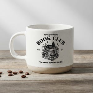 May include: A white ceramic mug with a tan base and a large handle. The mug features the text "HOUSE OF WIND BOOK CLUB" and a graphic of stacked books and a mountain. The words "VALKYRIE READING ROOM" are also printed on the mug.