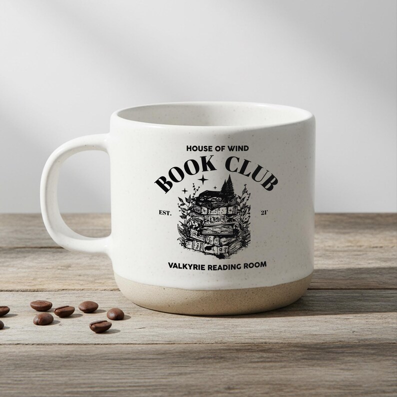 May include: A white ceramic mug with a tan base and a large handle. The mug features the text "HOUSE OF WIND BOOK CLUB" and a graphic of stacked books and a mountain. The words "VALKYRIE READING ROOM" are also printed on the mug.