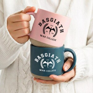 May include: A stack of two ceramic mugs with a matte finish. The top mug is light pink with black text reading BASGIATH WAR COLLEGE and a dragon graphic. The bottom mug is dark teal with white text reading BASGIATH WAR COLLEGE and a dragon graphic.