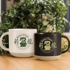May include: Two ceramic mugs, one white and one black, with a beige base. Each mug features a green circular logo with a dragon design and the text "Jasmine Dragon, The Finest Teas in Ba Sing Se."