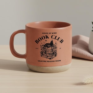 May include: A speckled, peach-colored ceramic mug with a cream-colored base. The mug features the text "BOOK CLUB" and "VALKYRIE READING ROOM" with a graphic of a stack of books and a house.