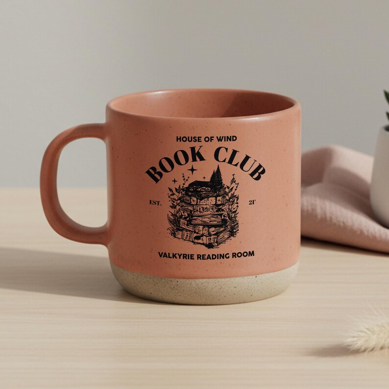May include: A speckled, peach-colored ceramic mug with a cream-colored base. The mug features the text "BOOK CLUB" and "VALKYRIE READING ROOM" with a graphic of a stack of books and a house.