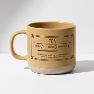 May include: A beige ceramic mug with a light beige base. The mug has the word "TEA" in a rectangular frame, with the words "Energy", "Comfort", and "Productivity" and corresponding numbers. The text "Will increase health and mood but will also result in frequent bathroom trips" is also present.