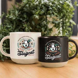May include: Two ceramic coffee mugs, one cream and one black, with a two-tone design. Each mug features a whimsical illustration of a slug-like creature in a coffee cup, with the text "The Slugcat Coffee". The mugs have a rounded base.
