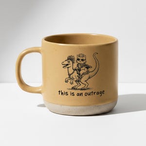 Könnte beinhalten: Senffarbene Keramik-Tasse mit cremefarbenem Boden. Die Tasse zeigt eine schwarze Illustration einer Katze mit Krone, die auf einem Dinosaurier reitet, mit dem Text "this is an outrage" darunter.