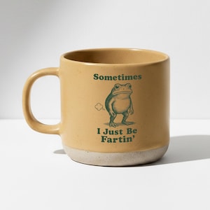 Op de afbeelding: Een bruin keramische mok met een groene kikker illustratie en de tekst "Sometimes I Just Be Fartin'". De mok heeft een afgeronde vorm met een handvat en een gespikkelde, gebroken witte basis.