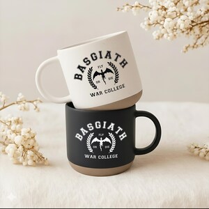May include: Two ceramic mugs stacked vertically. The top mug is off-white with black text reading Basgiath War College and a dragon graphic. The bottom mug is black with white text reading Basgiath War College and a dragon graphic. Both have tan bases.