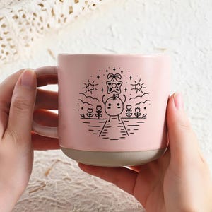 Peut inclure: Une tasse en c&eacute;ramique rose avec une base et une anse gris clair. La tasse pr&eacute;sente un dessin au trait noir d'un personnage de fruit souriant, entour&eacute; d'&eacute;toiles, de nuages et de fleurs. La tasse est tenue par deux mains.