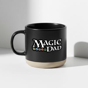 May include: Black ceramic mug with a beige base and handle. The mug features the text "MAGIC DAD" in white, with colorful symbols below the text. The mug is designed for hot beverages.