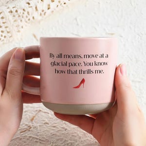 May include: A pink ceramic mug with a light gray base and handle. The mug features the text "By all means, move at a glacial pace. You know how that thrills me." and a red high heel illustration.