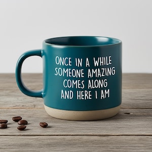 Pode incluir: Caneca de café de cerâmica azul-petróleo com o texto "ONCE IN A WHILE SOMEONE AMAZING COMES ALONG AND HERE I AM" em branco. A parte inferior da caneca é bege claro. Vários grãos de café estão espalhados na superfície de madeira.