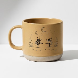 Może przedstawiać: Beżowy ceramiczny kubek z białą podstawą. Kubek przedstawia fantazyjną ilustrację tańczących owadów pod księżycem i gwiazdami. Uchwyt znajduje się po lewej stronie.