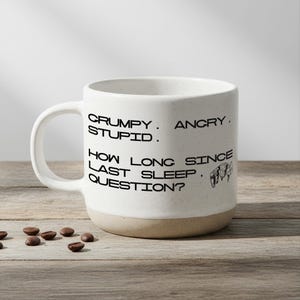 Könnte beinhalten: Weißer Keramikbecher mit schwarzem Text: "Grumpy. Angry. Stupid. How long since last sleep, question?" Der Becher hat einen beigen Boden und einen gebogenen Henkel. Kaffeebohnen liegen auf der Holzoberfläche verstreut.